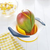 A ripe mango is being sliced in a mango splitter with black handles on a white table. A bowl of diced mango and a yellow gingham towel are in the background.