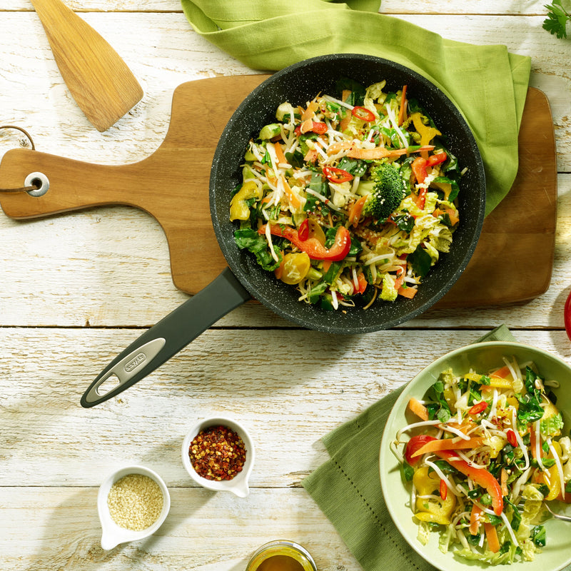 The Zyliss Ultimate Non-Stick Frying Pan holds colorful stir-fried vegetables on a wooden board beside a serving plate, bowls of sesame seeds and chili flakes, all set on a white table with a green napkin. #20cm/8"