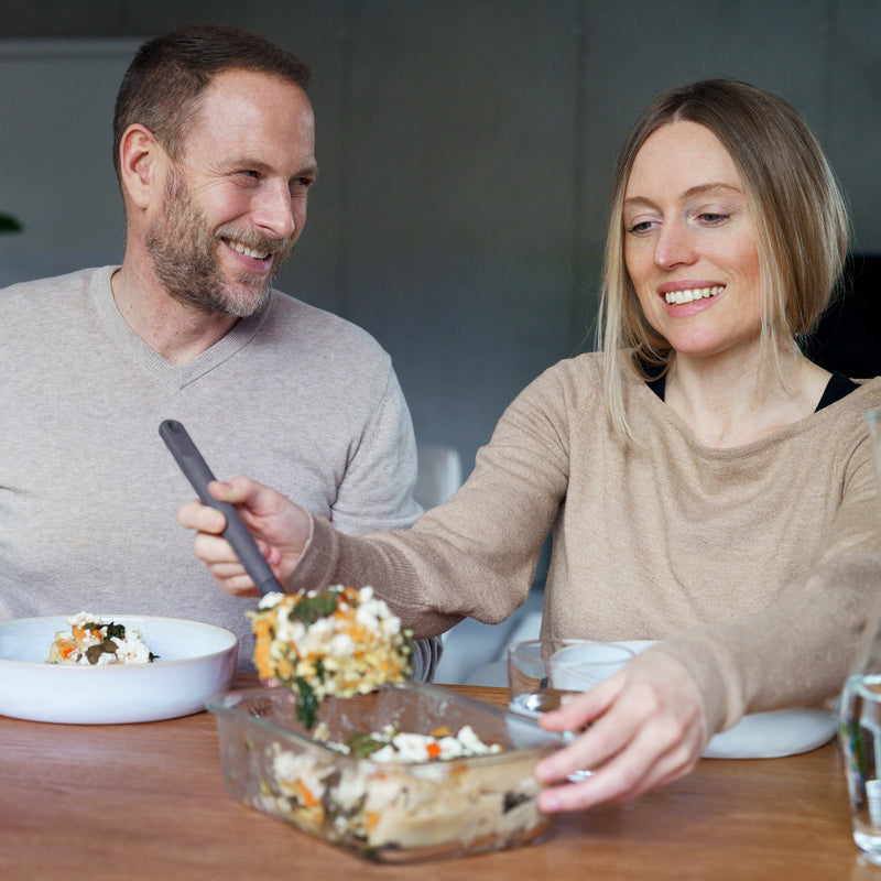 A smiling woman serves a casserole at a wooden table, with Zyliss Glass Containers nearby for easy meal prep. She and the man beside her look relaxed, enjoying a cozy indoor meal together. #0.37L/0.39qt
