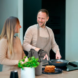 A man in an apron cooks at the stove, smiling at a woman beside him in their modern kitchen. Pancakes rest on the counter next to ergonomic utensils and a heat-resistant Zyliss Ladle, ideal for their morning routine. #Medium